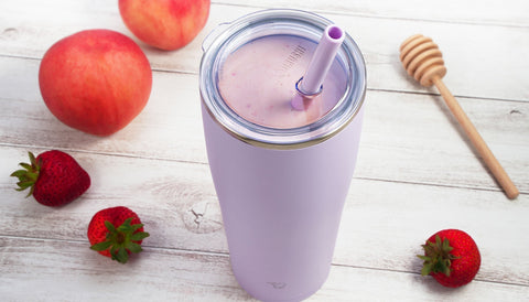 Strawberry and peach smoothie in a pastel purple Zojirushi insulated tumbler with straw, surrounded by fresh strawberries, peaches, and honey on a light wooden surface.