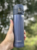 Hand holding a Zojirushi SM-KHE dark blue travel mug with flip-top lid and safety lock, outdoors background.
