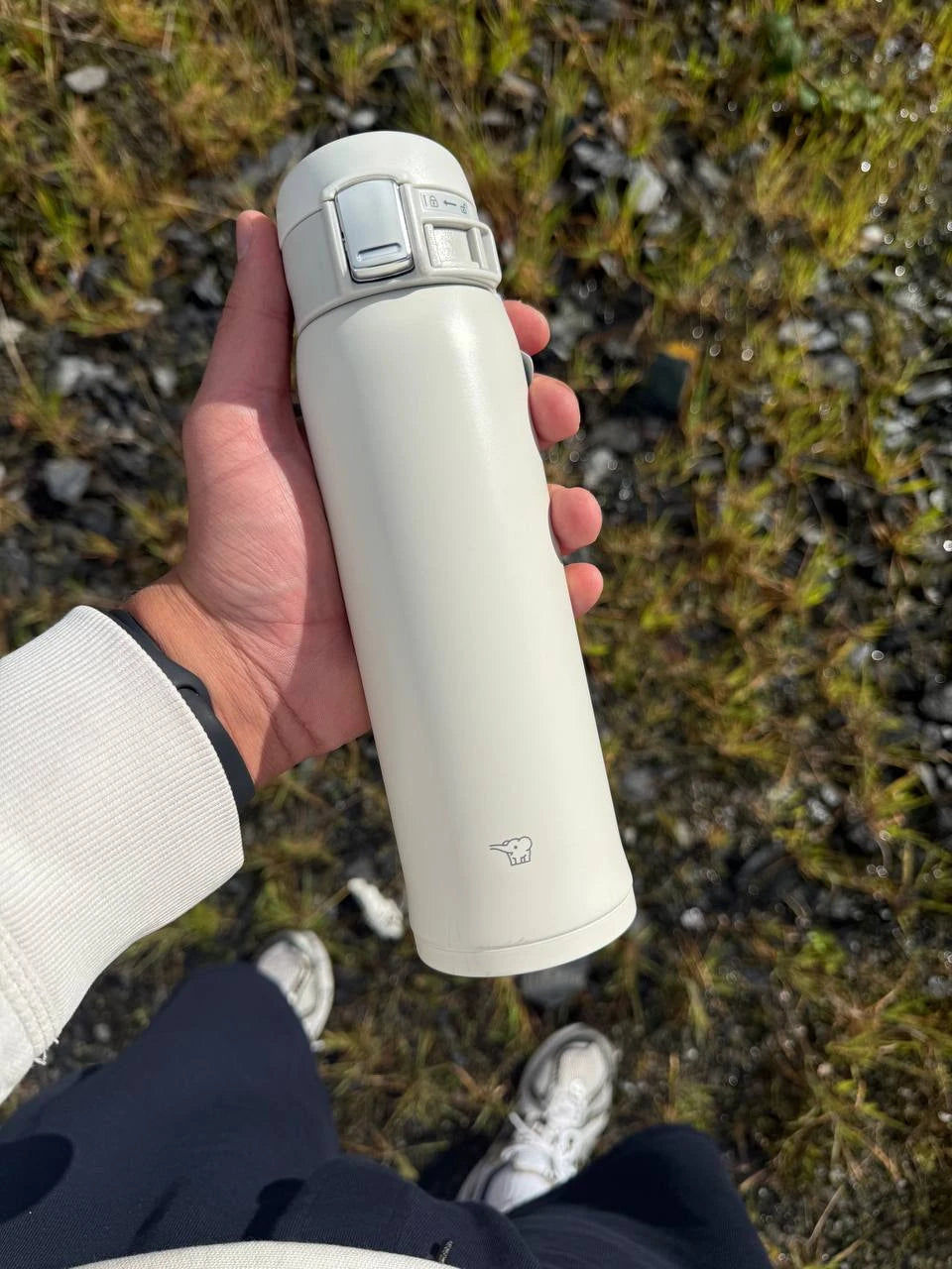 Hand holding a white Zojirushi Mug SM-SF (360–600 ml) with a one-touch locking lid, outdoors over grass and stones.