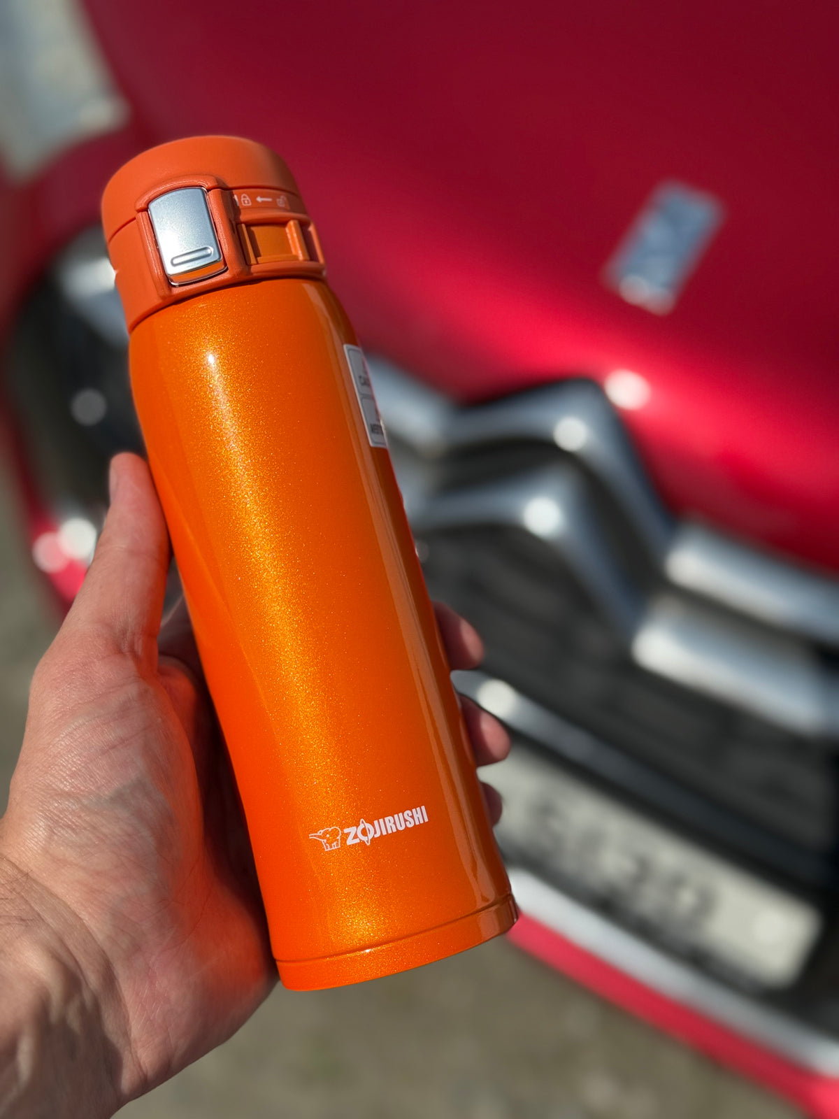 Zojirushi SM-SHE Orange travel mug held in hand with a red Citroën car in the background, 480 ml (16.20 oz) to 600 ml (20.30 oz)